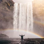 A man standing under a rainbow at a waterfall