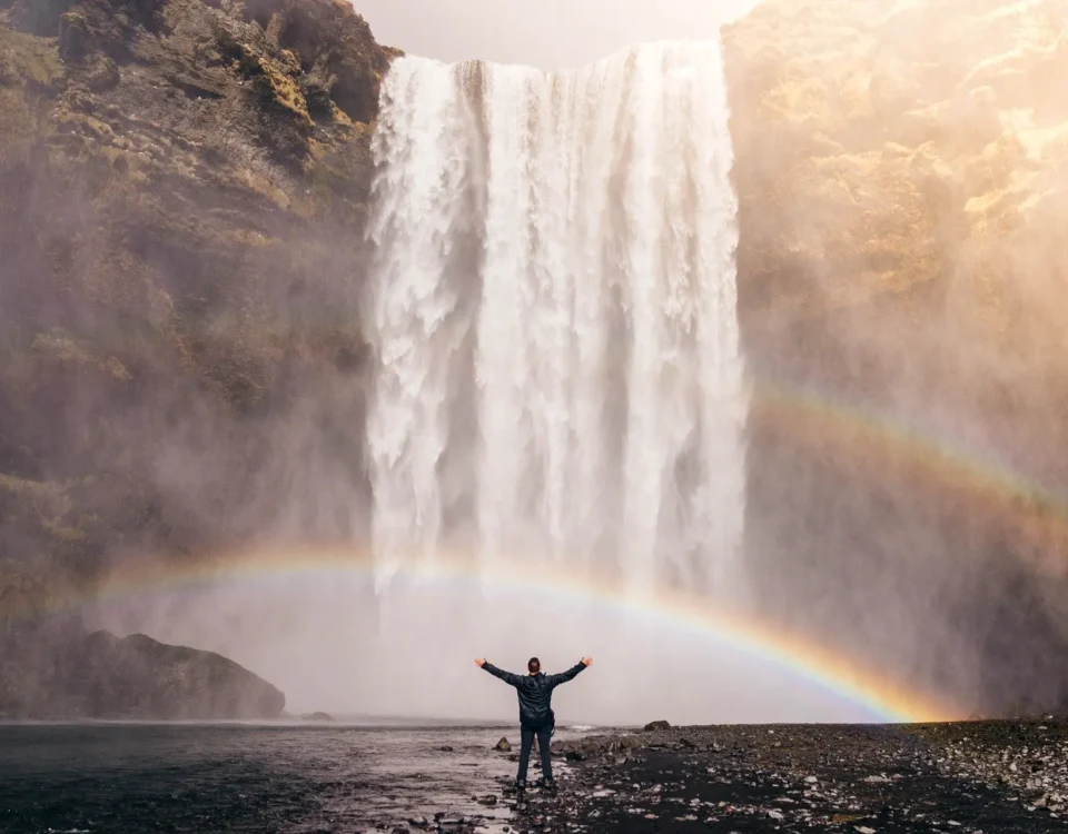 A man standing under a rainbow at a waterfall